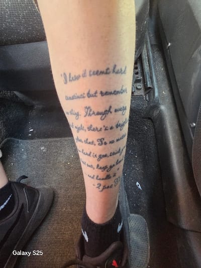 The image displays the lower leg of an individual, likely an adult, situated within the interior of a vehicle in Novo Delchevo, Bulgaria. The bare shin of the leg is extensively tattooed with black, handwritten script, which reads: "I know it seems hard sometimes but remember one thing. Through every dark night, there's a bright after that. So no matter how hard it gets, stick your heel out, keep ya head up and handle it. - 2Pac (R.I.P.)". The individual's foot is clad in a black athletic sock with a white logo visible near the ankle, and a dark-colored athletic shoe with laces and a thick sole. The immediate environment consists of a dark, possibly grey, carpeted floor on the left side of the frame, and a darker, textured plastic or rubber surface on the right. A ribbed pedal, consistent with a vehicle's accelerator or brake pedal, is partially visible at the bottom right. A section of a dark, textured fabric seat is visible in the upper left corner. The scene is illuminated by ambient light.