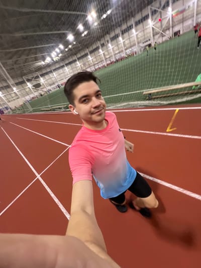The image displays a young man taking a selfie or being photographed on an indoor running track. The man is positioned in the foreground, looking directly at the camera with a slight smile. He has short dark hair and is wearing a bright pink t-shirt that transitions into a light blue or white gradient towards the bottom, black compression shorts or leggings, and light blue athletic shorts over them. He has white wireless earbuds in both ears. A portion of another person's arm or shoulder is visible in the bottom right corner, suggesting a selfie or close-up shot.
The setting is an indoor athletic facility. The man is standing on a reddish-brown running track with distinct white lane markings. Further down the track, a yellow arrow or symbol is visible. To the left and in the background, a large green artificial turf field, possibly for soccer or football, is visible, enclosed by a protective netting. The ceiling of the facility is high, with numerous bright overhead lights illuminating the space, and the structural beams are also visible. In the distance on the green field, several small, blurry figures can be seen, indicating other people are present at the facility. The man casts a shadow on the track to his right. The overall atmosphere suggests an active sports environment. The image is rotated 90 degrees clockwise.