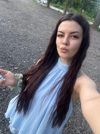 In the city of Zlati Voyvoda, Bulgaria, a young woman with long, dark brown hair and fair skin is captured in a selfie. She is wearing a light blue halter dress with a high neckline. Her expression is playful as she pouts her lips. Her right hand is visible, holding a light-colored object, possibly a phone, and adorned with a green scrunchie on her wrist. The background consists of a rough, gravelly surface, suggestive of a parking area or an unpaved road, with trees and greenery in the distance. A white car is partially visible on a road in the background, indicating a semi-rural or suburban setting within Zlati Voyvoda. The lighting suggests it is daytime, likely late afternoon or early evening given the soft illumination.