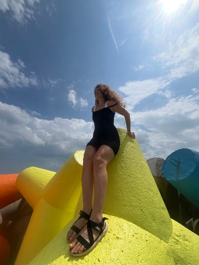 A young woman with curly brown hair, wearing a black dress and black sandals, sits on a bright yellow, multi-pronged concrete structure. She is positioned on the side of one of the prongs, facing to her left with her arms resting on the structure. The background consists of a bright blue sky with scattered white clouds, and the sun is visible in the upper right corner, creating lens flare. To the left of the yellow structure, a portion of an orange structure is visible, and to the right, a blue structure is partially seen. The concrete structures appear to be tetrapods or similar coastal defense units, commonly found along shorelines. The scene is outdoors, suggesting a coastal or harbor area. The image is captured from a low angle, emphasizing the sky and the size of the concrete structures. It is not possible to definitively determine the specific location within Phú Thủy, Vietnam, from the visual information provided.