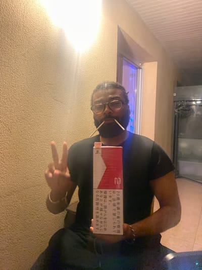 A man, with a dark complexion and wearing glasses, holds a pack of Marlboro cigarettes displaying Japanese text. He is also wearing a dark-colored shirt and is making a peace sign with his hand. The background features a wall with a textured surface, a window with a blue light visible inside, and some kind of metallic equipment. The location appears to be an indoor area, potentially within Montreuil, France.