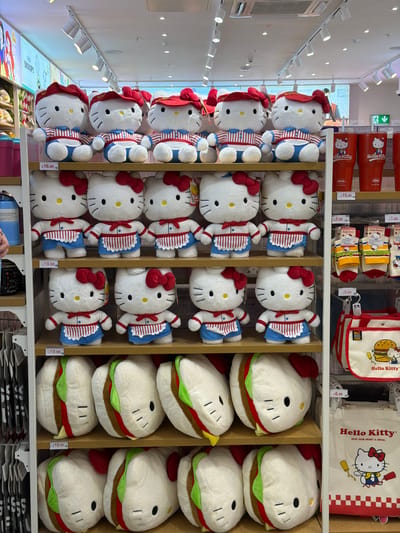A well-lit retail store shelf displays an assortment of Hello Kitty merchandise. The primary subjects are rows of plush toys and bags.
On the top shelf, three large Hello Kitty plush toys are seated, each wearing a red cap and a blue and white striped outfit with a red bow tie. Below them, another shelf holds six more Hello Kitty plush toys, similar in design but wearing red bows and blue overalls over striped tops.
The next shelf down features several large, round plush toys designed to resemble burgers with Hello Kitty faces. These are arranged horizontally. The shelf below that contains more of these burger-shaped Hello Kitty plush toys.
To the right of the plush toys, a display rack shows several items. The top section has three red tumblers featuring Hello Kitty. Below these are stacks of colorful socks, also with Hello Kitty designs, packaged in clear plastic. Further down, a white canvas tote bag with a Hello Kitty and burger graphic is visible. Below this, a larger white canvas tote bag displays a prominent Hello Kitty illustration with a slogan and a checkered red and white pattern at the bottom.
The shelves are constructed of light-colored wood, and the environment is brightly illuminated by overhead track lighting. Price tags are visible on some shelves. The overall presentation suggests a clean and organized retail setting within the city of Grays, United Kingdom.
