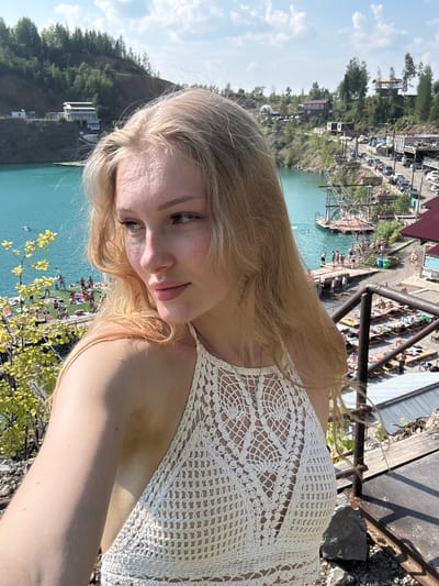 A young woman with blonde hair, wearing a white crochet top, poses for a selfie outdoors. She is positioned in the foreground, with her head turned to the left, looking away from the camera. The background reveals a scenic view of the Karyer Izvestnyak quarry in Russia. The quarry is filled with vibrant turquoise water, and its steep, rocky sides are covered with lush green trees. On the slopes and around the water's edge, various structures are visible, including buildings, platforms, and pathways, suggesting a recreational area. Several people are present, some sunbathing on loungers and others gathered near the water or on wooden decks. Cars are also visible in the distance, indicating roads and parking areas. The sky above is partly cloudy with a bright blue hue. The overall scene depicts a sunny day at a popular tourist destination.