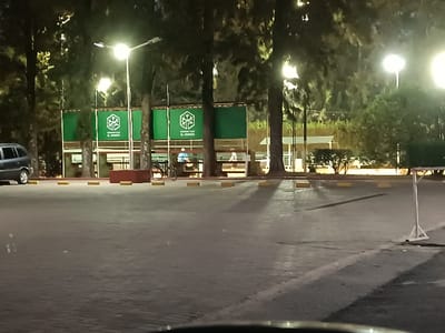 An outdoor parking area at night, illuminated by streetlights. A bus stop or covered seating area features green signage with the words "El Verdad" and a tree-like symbol. Sparse trees surround the area, with a few individuals visible seated within the bus stop. A parked car is on the left, while parking markers are laid out on the pavement. Shadowy areas intersperse with bright light across the paved surface.
