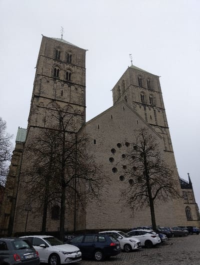 The image depicts the St. Paul's Cathedral (St. Paulus Dom) in Münster, Germany, under an overcast sky. The prominent westwerk of the Romanesque cathedral features two tall, square stone towers with multiple tiers of arched windows, each topped with a conical spire and a weather vane. The central section between the towers presents a large, unadorned light-colored stone facade, characterized by a symmetrical arrangement of twelve circular windows. To the left, a section of green-tiled roof is visible. Several bare deciduous trees stand in front of the cathedral, indicative of a colder season. In the foreground, a row of vehicles is parked on a cobblestone surface. From left to right, visible vehicles include a dark grey Fiat 500, a white Volkswagen Golf, a dark blue Volkswagen Golf Variant, a white Hyundai i20, and other less distinguishable cars extending into the distance along the building's side. No individuals are visible in the scene. The perspective is a low-angle shot, looking upwards at the cathedral.