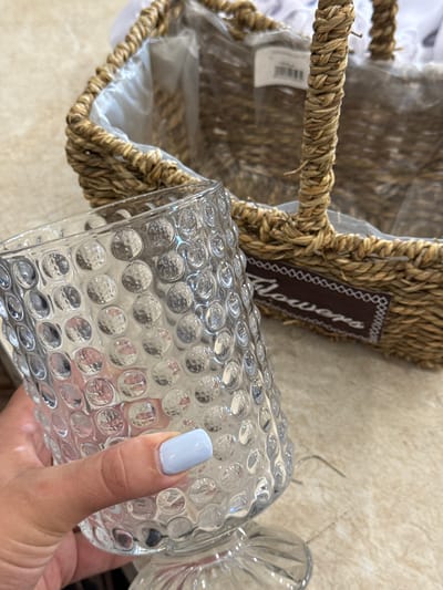 A close-up, eye-level shot shows a hand holding a clear, textured glass. The glass has a hobnail or bubble pattern and a pedestal base. A person's thumb and index finger are visible, with a light blue nail polish on the fingernail. In the background, a woven basket with a plastic liner is partially visible. A dark brown ribbon with white decorative stitching and the word "Flowers" is attached to the basket. The basket appears to be made of natural fibers. The setting is indoors, with a light-colored, textured surface beneath the objects. The image does not contain any information about the city of Sunzha, Russia.