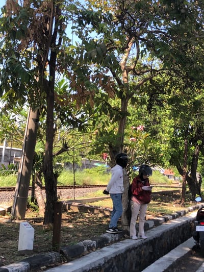 Two individuals stand near a set of railroad tracks in Buduran, Indonesia, beneath a canopy of trees. The individual on the left wears a white hoodie and blue jeans, while the individual on the right is dressed in a maroon jacket and light-colored pants. Both are wearing helmets, suggesting they may have arrived by motorcycle, one of which is partially visible in the foreground. The environment is rural, characterized by lush green grass and dense foliage. A white marker with the inscription "KAI PT. KAI" is visible in the lower left corner, indicating the presence of Indonesian railways. The scene is bathed in daylight, with the sun casting shadows on the ground.