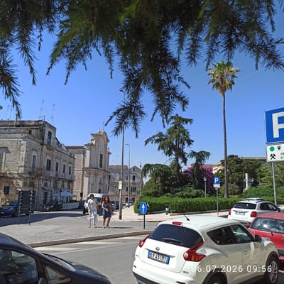 In Ruvo di Puglia, Italy, on July 6, 2026, at 09:56, a street scene unfolds under a clear blue sky. The foreground is partially obscured by the dark green needles of pine trees. A white Nissan Juke is parked in the lower right corner, its license plate reading "EV 431MY". To its left, the front of a dark-colored car is visible.
In the mid-ground, a cobblestone street crosses the frame, bordered by a sidewalk. Two women are walking on the sidewalk, one in a white dress and the other in a blue dress. Behind them, a white van is parked. Further back, a dark sedan is parked on the street. Across the street, a row of stone buildings with arched windows and balconies lines the road. A church with a domed roof and a light-colored facade stands prominently in the background.
The environment is characterized by Mediterranean architecture and lush greenery, including palm trees and manicured hedges. Traffic signs are visible, including a blue parking sign with a white "P" and a sign with a white arrow pointing upwards on a blue circular background. The overall impression is of a sunny day in an Italian town.