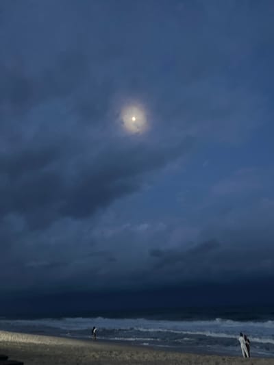 The image depicts a beach scene at dusk, with a full moon visible through scattered clouds. The sky is a deep blue, with darker, heavier clouds dominating the lower half of the frame and lighter, wispy clouds around the moon. The moon is a bright, luminous orb, casting a faint glow. Below the sky, the ocean is visible with gentle waves breaking on the shore. The beach itself is sandy and appears to be unoccupied except for two small figures. On the left side of the image, a single person is standing on the wet sand near the water's edge, facing the ocean. On the right side of the image, two figures, appearing to be a couple, are standing together on the sand, also facing the ocean. The overall impression is one of a tranquil evening by the sea, with the moon providing a soft illumination. The location is identified as Mopo-Akinlade, Nigeria, a coastal community known for its beaches.