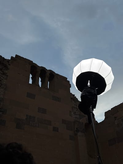The image captures a low-angle view of a building in Anipemza, Armenia, taken in daylight. Dominating the upper part of the image is a bright, lantern-like light diffuser held in place by a dark arm extending from the building. The building itself is constructed from stone blocks, with a series of three openings or windows visible in the structure. The sky is visible through the openings and around the structures, revealing a cloudy day. A small portion of a person or object is visible in the bottom right corner.