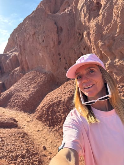 The image, when correctly oriented, captures a smiling woman against a backdrop of distinctive reddish-brown rock formations. The subject is a woman with long, straight blonde hair and blue eyes, looking directly at the camera with a wide, cheerful smile. She is wearing a light pink long-sleeved crew-neck shirt and a matching pink baseball cap. White-framed sunglasses are perched on top of her head. A subtle, light-colored design, possibly a star or abstract shape, is visible on the front of her cap, but no clear text is legible.
The setting is an outdoor, natural environment, characterized by large, rugged rock formations that exhibit a rich, reddish-brown hue and varied textures. These geological features are consistent with landscapes found in Tosor, Kyrgyzstan, known for its red rock canyons. The bright sunlight suggests it is daytime, casting shadows that emphasize the contours and depth of the rocks. A small section of clear blue sky is visible in the lower left corner, indicating fair weather. The woman's joyful expression and the scenic natural background convey a sense of enjoyment and appreciation for the outdoor surroundings. No additional discernible text is visible in the image.