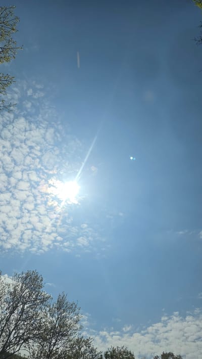 The image shows a clear, blue sky with scattered altocumulus clouds and the sun shining brightly, creating a lens flare effect. Tree branches are visible along the edges of the frame, suggesting the image was taken from a ground-level perspective. The location, Stara Kresna, Bulgaria, cannot be independently verified from the image content alone.