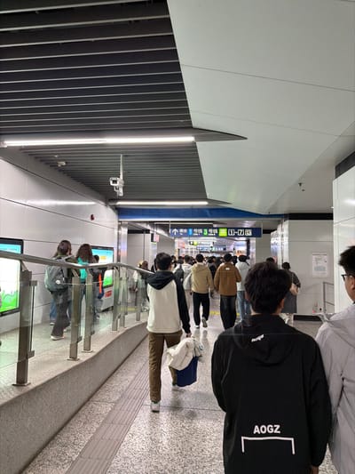 An indoor scene depicts a brightly lit, modern subway station concourse or passageway in Yuhuatai District, China. The environment features a dark gray slatted ceiling with linear white fluorescent or LED light fixtures, and white flat panel walls. A white security camera is mounted overhead on the left. The floor is composed of light gray terrazzo-style tiles, with tactile paving strips visible in the foreground.
Approximately 20-30 individuals, predominantly young adults, are present. Most are observed walking away from the camera towards a junction in the midground, suggesting movement towards platforms or exits. In the immediate foreground, the back of a person wearing a black hoodie with "AOGZ" printed in white on the back is visible on the right. To their left, another person, also seen from behind, wears a white and black hoodie, brown trousers, and white sneakers, carrying a blue bag containing white fabric in their left hand.
On the left side of the scene, a raised platform is separated from the main walkway by a glass barrier with stainless steel posts and a top rail. Behind this barrier, several individuals are standing or walking. Digital screens displaying green graphics are embedded in the wall behind them, partially obscured. A red fire alarm pull station is mounted on the left wall. In the midground, a prominent blue overhead sign with white text and arrows directs passengers to "Line 1" and "Line 3," and indicates exits "1-7" in both Chinese and English. People in the background are densely clustered, navigating the passageway. No direct interactions between individuals are discernible; all subjects appear to be in transit.