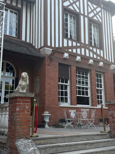 The image shows the exterior of a brick building with a timber-framed upper story, characteristic of Norman architecture, likely located in Saint-Vaast-du-Val, France. The building features multiple tall, multi-paned windows with white frames. A statue of a dog, possibly a Basset Hound, sits on a brick pillar to the left of the entrance. In front of the building, a set of stone steps leads up to a patio area where a white metal table and four chairs are arranged. Red velvet ropes on gold stanchions are present, suggesting a controlled entrance or a VIP area. The overall scene appears to be a daytime shot, with natural light illuminating the facade.