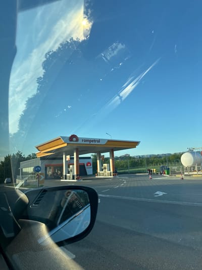 This is a wide-angle photograph taken from inside a vehicle, looking out through the windshield and side mirror towards a Rompetrol gas station. The sky is a clear, bright blue with some wispy white clouds. The sun is visible, high and slightly to the left, casting a bright glare on the windshield.

The gas station features a prominent yellow and white canopy covering multiple fuel pumps. The Rompetrol logo, a red flower-like symbol, is visible on the canopy and on a building behind the pumps. A large white cylindrical tank, likely for storing fuel, is located to the right of the station. The ground is paved with interlocking grey bricks, and white directional arrows are painted on the asphalt. A few traffic cones are placed near the pumps. In the background, there are trees and a distant row of buildings.

The side mirror reflects the interior of the car, showing a portion of the dashboard and a glimpse of the driver. The reflection also captures part of the gas station and the sky. The overall impression is a sunny day at a modern gas station, likely located on the outskirts of a city. The image appears to have been taken in Trușeni, Moldova, given the context provided in the prompt.