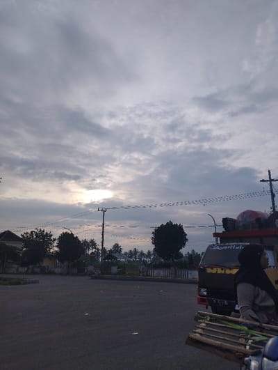 The image captures an outdoor scene under an overcast sky, likely during dawn or dusk, given the soft, diffused light breaking through the clouds. The setting appears to be a roadside or a parking area in a rural or semi-urban environment in Kecamatan Jangkar, Indonesia.
In the foreground on the right, a person wearing a black hijab and a light-colored long-sleeved top is visible from the side, looking towards the right. They appear to be seated or standing next to a yellow and brown truck, the front portion of which is partially visible. The truck has items loaded on its bed, suggesting it might be a cargo or utility vehicle. Faintly visible on the truck's license plate is the text "P 9338 HG". In the extreme lower right foreground, a stack of wooden planks or slats is partially visible.
The midground features an asphalt surface leading into the distance. Power poles with numerous overhead power lines stretch across the frame, creating linear patterns against the sky. Beyond the road, a line of lush green trees, including palm trees, and some distant low-rise buildings are visible. The sun is a bright spot partially obscured by the clouds in the upper-left midground. The overall mood is subdued, with no clear activity taking place other than the presence of the person and vehicles.