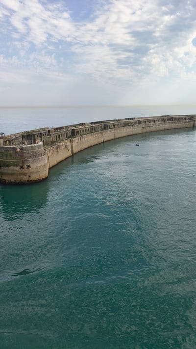 The image captures a broad view of the Dover Patrols war memorial in Dover, United Kingdom. The memorial, a long, curved concrete structure with multiple openings resembling gun emplacements, extends into the tranquil, teal-colored water. Seagulls are visible perched on top of the structure, and a small boat with two occupants is present in the water to the right of the memorial. The sky above is a pale blue, adorned with scattered, wispy white clouds. The overall scene suggests a calm, overcast day with a muted color palette.