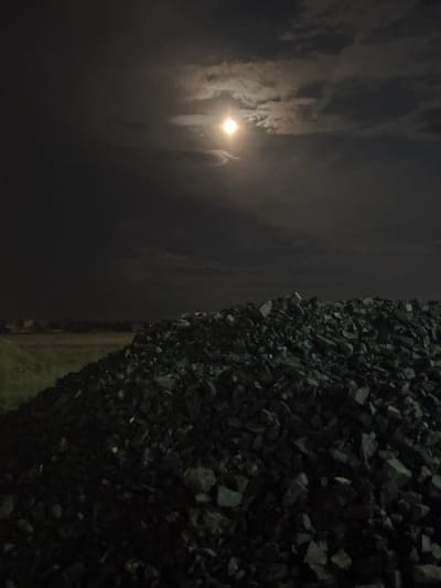 The image displays a nocturnal scene featuring a bright moon partially obscured by clouds, juxtaposed with a large pile of dark, angular rocks.
On the right side of the frame, a glowing orb, presumably the moon, is prominently visible against a dark night sky. It shines brightly through a veil of scattered clouds, which vary in density—some appearing thin and wispy, others more substantial. This celestial element dominates the upper portion of the frame, creating a moody and atmospheric impression.
The left side of the image is occupied by a dense mass of dark, irregularly shaped rocks or gravel. This pile extends vertically, creating a rough, textured surface. The rocks are uniformly dark, with subtle variations in light and shadow indicating their jagged forms.
A faint, lighter strip, possibly a path or ground, is discernible along the bottom edge where the rock pile meets the darker expanse of the background. No people or specific activities are visible. The time of day is clearly night, indicated by the moon and dark sky. There is no visible text in the image.