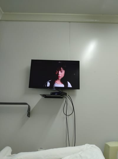 The image shows a television screen displaying a close-up of a young Asian female's face. She has dark hair and is wearing a light-colored top. The TV is mounted on a white wall, above a black shelf holding a small electronic device with cables. The wall appears to be made of panels and has some subtle lighting effects. There is a metal bracket mounted on the wall to the left of the television, and a white sheet or blanket is visible at the bottom of the frame, suggesting a bedroom or hotel room. A yellow object, possibly a small appliance or furniture, is partially visible on the right. The scene is dimly lit, with a spotlight effect on the wall to the right. The inscription "220 V" is visible on the wall near the bottom right. The image is taken indoors in what appears to be a sparsely decorated room. The location is identified as Қопа ауылдық округі, Kazakhstan.