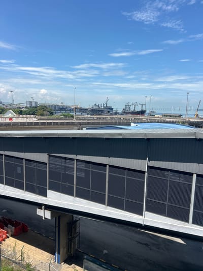 This is an eye-level, medium shot captured on a sunny day, looking across a modern grey overpass with dark, slatted windows. The overpass extends into the background, bridging over an asphalt road with yellow markings. In the distance, beyond the overpass, lies a bustling port area. Several large cargo ships are docked, and numerous cranes are visible, indicating active maritime operations. Silos and industrial buildings are scattered throughout the port. A long bridge, possibly a highway or railway bridge, spans across the water in the far background, connecting to land on the right side of the image. The sky is a vibrant blue with scattered white clouds. To the left of the overpass, red and white construction barriers are stacked on the ground. The scene appears to be located in Butterworth, Malaysia, a city known for its industrial and port activities.