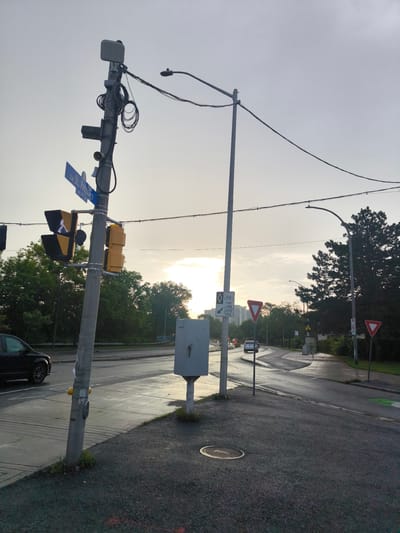 This is a street-level, eye-level, medium shot of an intersection with a traffic light and street signs. The sky is overcast and appears to be dawn or dusk. The road is wet, suggesting recent rain. A black SUV is visible on the left side of the road.
On the right side of the image, a concrete sidewalk curves away from the road. There are several street signs visible, including a yield sign, a sign indicating "THIS LANE / C'ETTE VOIE" with a bicycle symbol, and another yield sign. A bus shelter with a bench is also present. Trees line the background.
The traffic light on the left has a pedestrian signal displaying a white walking person symbol. A street sign reads "rue St. Patrick St." The traffic light pole is also equipped with various wires and antennas, suggesting it is a hub for communication equipment. A grey electrical box is situated on the right side of the road.
The overall scene appears to be an intersection in a suburban or residential area. Based on the street sign "rue St. Patrick St.", this intersection is likely located in Ottawa, Canada.