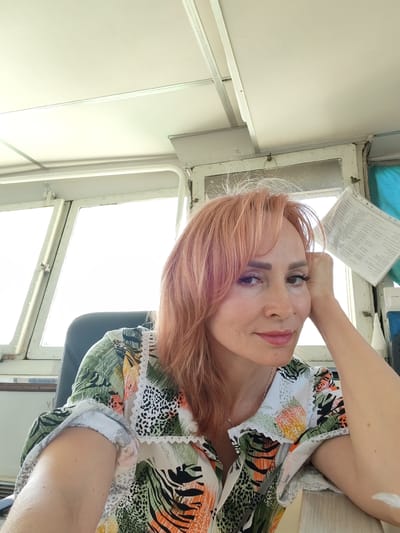 A woman with reddish-blonde hair and light skin is pictured indoors, likely in the cabin of a vessel due to the large windows and visible nautical equipment. She is seated, leaning her head on her hand, and looking towards the camera with a slight smile. She is wearing a colorful, patterned short-sleeved shirt. The background shows large, multi-pane windows that appear to be letting in bright daylight, suggesting it is daytime. There is also a document clipped to the window, showing lines of text which are not legible. The overall mood seems relaxed and contemplative.