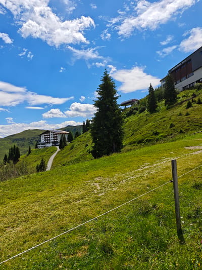 The image depicts a mountainous landscape in Kirchberg in Tirol, Austria, under a partly cloudy blue sky. In the mid-ground, a large, white hotel with a brown roof and solar panels is situated on a grassy hillside. A winding dirt road leads up to the hotel. To the right of the hotel, a tall, dark green evergreen tree stands prominently. Further up the slope, a modern building with a dark facade and the text "FLECKALMBAHN 1800m" is visible, suggesting a cable car station. Another, smaller building with a red roof and a covered structure is also present on the hillside in the upper left quadrant. The foreground consists of a green, grassy field with scattered wildflowers and a white rope fence supported by a wooden post. The overall scene conveys a tranquil, alpine environment.