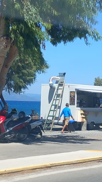 A person wearing a blue shirt and tan shorts walks to the right, carrying a blue bucket and a black bucket. The person is in front of a white food truck with a closed window. A green ladder leans against the food truck, with a silver ventilation pipe extending from the roof and curving over the ladder. In the background, the sea is visible under a clear blue sky, with hills in the distance. To the left of the food truck, two motorcycles are parked. The scene appears to be near the coast, possibly in a town or city.