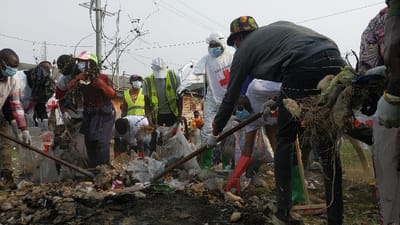 The image captures an outdoor community cleanup operation in Gisenyi, Rwanda, focused on clearing a substantial accumulation of refuse on unpaved ground. Approximately ten individuals are actively engaged in removing a heterogeneous mound of waste consisting of plastic bags, organic debris, fabric scraps, and various unidentifiable items. Most participants wear surgical face masks and protective gloves. Several individuals are affiliated with the Red Cross/Red Crescent, identified by branded white clothing, including one person in a full white hazmat-style suit displaying "CROIX-ROUGE" and a red cross emblem. At least two individuals are wearing bright yellow reflective vests. Personnel are using long-handled brooms and rakes to gather the trash, which is being collected into transparent plastic bags. The background includes overhead power lines, a prominent metal utility pole with multiple wires, distant wooden structures, and a partial barbed wire fence on the right. The sky appears overcast or hazy.