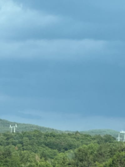 The image depicts a rural landscape under a cloudy sky. The foreground is dominated by lush green trees, suggesting a natural or forested area. In the middle ground, rolling hills covered in trees extend across the frame. On the crest of one of the hills, a tall white structure, possibly a transmission tower or pylon, is visible. The sky is a deep blue with layers of clouds, hinting at overcast or potentially stormy weather. The overall impression is one of a natural, undeveloped area with some signs of human infrastructure. The location is identified as Dolno Varshilo, Bulgaria.