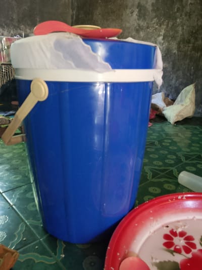 A blue plastic bucket with a white rim and a beige handle is on a green patterned floor in Merbopuntong, Indonesia. A white cloth is draped over the top of the bucket, and a red spatula is resting on top of the cloth. A red and white plate with a floral design is in the foreground, to the right of the bucket. A pale green object, possibly a bottle or container, is also visible to the right of the bucket. The background shows a textured, grey wall and some scattered items, including a light-colored bag. The scene appears to be indoors, possibly in a kitchen or storage area.