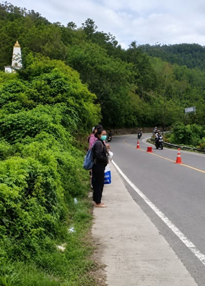 This image depicts a roadside scene in Sosor Dolok, Indonesia. In the foreground, a woman wearing a light blue face mask and a dark jacket stands on a concrete sidewalk next to a dense green bush. She is carrying a blue backpack and a white shopping bag. To her left, partially obscured by the foliage, are two other individuals, one wearing a pink top. A white structure with gold accents, possibly a monument or a decorative pillar, is visible behind the bushes on the left. The background features a winding asphalt road with a yellow line marking its edge. Several motorcyclists are traveling on the road, and orange traffic cones are placed along the roadside. The road is bordered by lush green trees and vegetation climbing up a hillside under a cloudy sky.