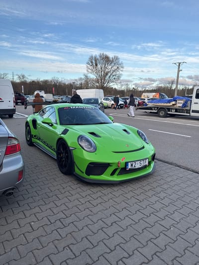 This image depicts a daylight scene in a paved parking area in Wolica, Poland. The primary subject is a bright lime green Porsche 911 GT3 RS MR, prominently parked and angled towards the left. The vehicle features black multi-spoke wheels, a large rear wing, and a front splitter with a visible red tow strap. A "Manthey" banner is displayed on its windshield, and "GT3RS MR" decals are present on its side skirts. The front license plate reads "W2 STF", indicating a Polish registration.

To the immediate left of the Porsche, the rear section of a silver sedan is partially visible. In the background, numerous vehicles are parked across the grey interlocking paver lot, including a white panel van, a dark SUV, and various other cars. Several individuals are present in the background; one person in a brown hooded jacket stands near the rear of the green Porsche, while others are seen walking or standing further back among the parked vehicles. On the right, a white flatbed tow truck is parked, carrying a load covered by a blue tarp and secured with wooden pallets. An individual in white pants and a dark top is seen walking past this tow truck. The background features a treeline of bare deciduous trees, with a prominent large, leafless tree in the center. Utility poles and overhead power lines are visible against a partly cloudy sky. The paved area transitions into an asphalt road on the right side of the frame.