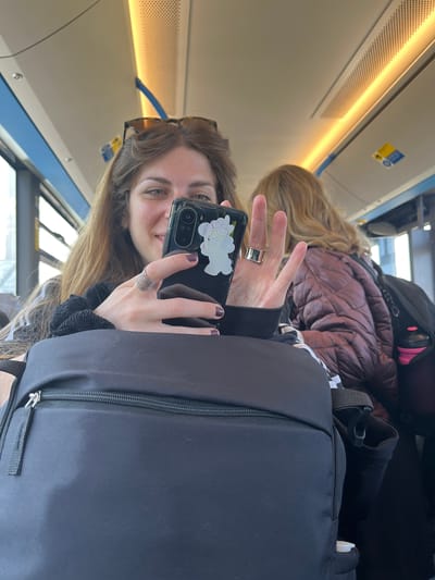 A selfie on a train in Vecsés, Hungary, shows a young woman holding a phone decorated with cloud stickers. She has long brown hair and wears sunglasses atop her head, gesturing with her hand that has rings. In the background is another woman with long blonde hair wearing a puffy maroon jacket and a black backpack. The interior of the train is visible, including the overhead lighting and window views.