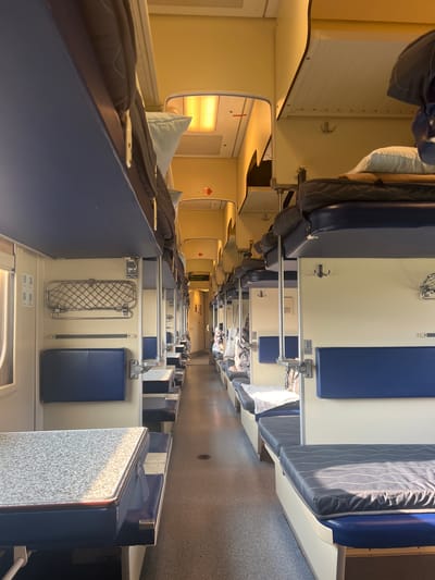 The image shows the interior of a train car, likely a sleeper carriage, with bunks arranged along the sides. The scene is brightly lit, suggesting it is daytime. The bunks are covered with dark striped bedding and light blue pillows. Blue fabric panels are visible on the upper bunks. Along the walls, there are various features typical of a train, such as hooks, luggage racks, and small tables. Some areas appear to have yellow walls. The overall impression is one of order and utility, with the bunks suggesting sleeping accommodations for passengers. The setting appears to be in Russia, specifically near Pugachevo. There is no clear indication of people being present, but the bunks are prepared for use. The lighting casts a warm glow throughout the carriage.