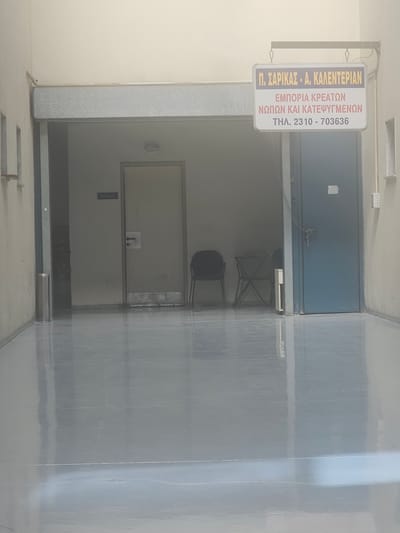 The image shows the entrance to a building in Menemeni Municipal Unit, Greece. A sign is visible that reads "Π. ΣΑΡΙΚΑΣ - Α. ΚΑΛΕΝΤΕΡΙΑΝ ΕΜΠΟΡΙΑ ΚΡΕΑΤΩΝ ΝΩΠΩΝ ΚΑΙ ΚΑΤΕΨΥΓΜΕΝΩΝ ΤΗΛ. 2310 - 703636". This translates to "P. SARIKAS - A. KALENTERIAN MEAT TRADE FRESH AND FROZEN TEL. 2310 - 703636". The entrance leads to a corridor with doors and chairs. The floor is reflective and appears to be a polished concrete or epoxy finish. The overall environment suggests a commercial establishment, possibly a butcher shop or a food processing business.
