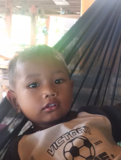 A close-up, slightly blurry selfie-style shot shows a young child lying in a dark hammock. The child has dark hair and a light brown complexion. Their eyes are open and looking towards the camera. The child is wearing a light-colored t-shirt with a graphic design featuring a soccer ball, the word "VICTORY," and the number "1970." The background of the image is out of focus, showing wooden structures and some greenery, suggesting an outdoor or semi-outdoor setting. The scene appears to be located in Pompanua, Indonesia, based on the context provided.