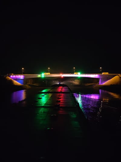 The image captures a night scene featuring a bridge illuminated with colorful lights. The bridge spans a body of water, reflecting the lights and creating a vibrant display. The bridge has white lights on top of the green and red color. The scene appears to be in Podosinki, Russia, given the context. The illumination suggests it may be a decorative or celebratory lighting scheme. The foreground includes a textured surface, partially submerged in water. The bridge also has 2 yellow vehicles crossing it. The overall environment is dark, allowing the colored lights to stand out.