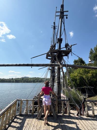 A woman with her back to the camera stands at the helm of a wooden ship, holding onto the wheel. She is wearing a pink t-shirt and patterned shorts, with a straw hat on her head. The ship appears to be docked on a body of water, with a tree-lined shore and a hill in the distance. The sky is blue with scattered clouds. The ship's rigging and wooden structure are prominent in the foreground. The scene is set during the day, likely in summer, given the foliage. The specific location in Satka, Russia, is a tourist attraction, likely a replica of a historical ship.