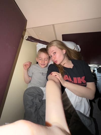 A Caucasian woman with long blonde hair and dark red painted nails lies next to a young Caucasian boy with blonde hair in what appears to be a sleeping compartment on a train. The woman is wearing a black t-shirt with red text. The boy is wearing a gray t-shirt with a graphic design. They are both lying on a bed with white bedding and a gray blanket. The interior of the compartment consists of brown walls and panels. The location is possibly Bobrov, Russia. The woman is holding her arm out as if taking a selfie.