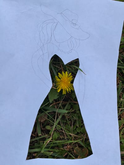 The image shows a piece of white paper with a dress silhouette cut out of it, placed on a patch of grass. A yellow dandelion is visible through the cut-out portion of the dress. A sketch of a woman's upper body, including a hat and necklace, is drawn on the paper. The grass appears green with some dry leaves and twigs scattered throughout. The lighting suggests an outdoor setting, possibly during the daytime. There is no indication of a specific location in Sterlitamak, Russia.