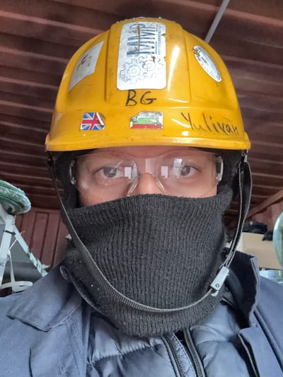 A close-up, frontal portrait captured in Hook of Holland, Netherlands, depicts an individual wearing personal protective equipment in an industrial or work setting. The person wears a yellow hard hat featuring a "KAMP Manufacturing Company" sticker, a small Union Jack flag sticker, a small Bulgarian flag sticker, and handwritten identifiers "BG" and "Yulivan." Clear safety glasses cover the eyes, which are the only visible facial features. A dark grey knitted neck gaiter covers the lower half of the face and neck. A dark strap with a light-colored clip is visible across the neck gaiter. The individual's attire includes a dark blue or navy collared work jacket over a dark puffer jacket. The blurred background consists of reddish-brown surfaces, potentially corrugated metal, and a partial view of a white and green structure on the left.