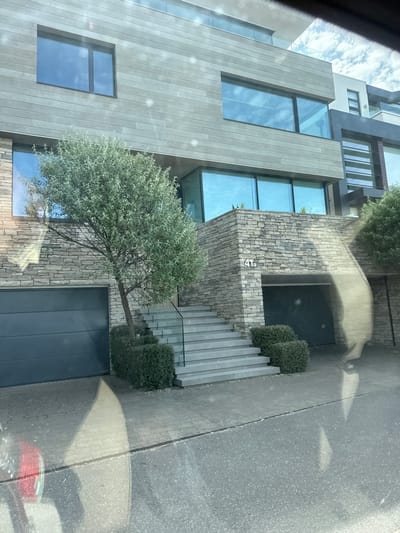The image shows a modern, multi-story residential building with a stone and wood facade. The building features large windows, a balcony with glass railings, and two garage doors. A paved walkway leads to a set of stone stairs that ascend to the entrance. A small tree and manicured bushes are positioned in front of the building. The address "41a" is visible on the stone facade. The scene is located in Poole, United Kingdom.