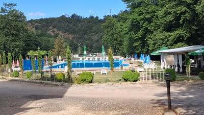 The image depicts an outdoor swimming pool facility in Ognyanovo, Bulgaria. The scene features a bright blue swimming pool, surrounded by white lounge chairs and colorful umbrellas. The pool area is set against a backdrop of lush green trees and a rolling hillside. A shaded seating area with white curtains and green umbrellas is visible to the right, possibly a cafe or bar. A gravel path leads to the foreground, with decorative bushes and cypress trees bordering the pool. The sky is clear and blue, indicating a sunny day. There are no people visible in the image.