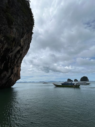 The image depicts a dramatic seascape with a large, dark rock formation dominating the left side of the frame. The rock face is rugged and textured, with some sparse vegetation clinging to its upper edges. The sea is a deep, rippling green, reflecting the overcast sky.

In the distance, several smaller, conical islands rise from the water, also covered in lush green foliage. A few boats are visible on the water. In the mid-ground, a long, green boat with a canopy is moored. Behind it, a white speedboat with an inscription that reads "J. CAROLING TOUR" is moving. Further out, other boats and islands can be faintly seen. The sky is filled with heavy, grey clouds, suggesting an impending rain or a generally moody atmosphere.

The scene is located in Tambon Krasom, Thailand, which is known for its karst topography and numerous islands in Phang Nga Bay. This particular view likely captures a scene within the bay, possibly near Ko Phi Phi or a similar island group famous for its towering limestone cliffs and emerald waters. There are no people clearly visible in the image, though the presence of the boats suggests tourist activity.