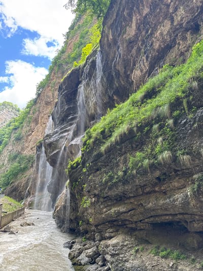 The image depicts a natural scene featuring a waterfall cascading down a rugged cliff face. A river flows at the base of the cliff, with rocks and vegetation dotting the landscape. The sky is partly cloudy, with patches of blue visible. The location is identified as Khushto-Syrt, Russia. The scene appears to be a natural landscape with no visible human activity or structures within the immediate vicinity of the waterfall and river. The focus is on the geological formations and the natural elements of the environment.