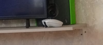 The image shows a white computer mouse on a shelf. Behind the mouse is a black speaker with a green border and a headphone attached to it. A portion of a dark blue computer screen is visible to the left of the speaker. The shelf is light brown and has some texture on it. The background appears to be a textured wall. The scene is indoors and likely in a home or office setting. There is no indication of the location in Dovzhansk, Ukraine.
