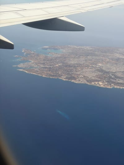 The image shows an aerial view from an airplane window, featuring a portion of the wing and a landscape below. The landscape appears to be coastal, with land mass densely covered by what seems to be urban development, extending along the coastline. A significant body of water occupies a large portion of the image. The geographical location is likely near Luqa, Malta, based on the urban layout and coastal features visible from the air. There are no people visible in the image.