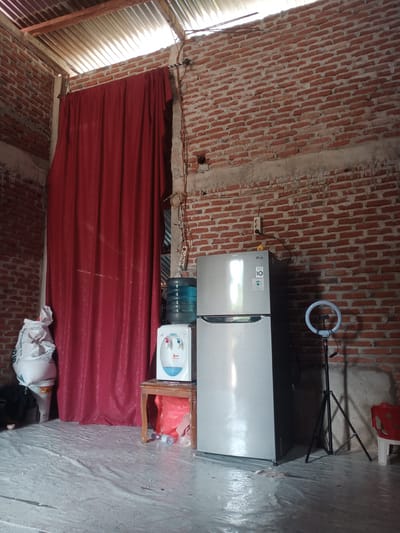 The image depicts an indoor scene with unfinished brick walls and a corrugated metal roof. In the center of the frame is a silver LG refrigerator. To the left of the refrigerator is a wooden stool topped with a water dispenser and a large blue water jug. A large white bag, possibly containing flour or other bulk goods, sits on the floor to the left of the stool and curtain. A large red curtain hangs on the left side of the image. To the right of the refrigerator, a black tripod with a circular ring light is set up. The floor is a textured, possibly concrete or tiled, surface. A small red plastic chair is visible on the far right. The scene appears to be in a utilitarian space, possibly a kitchen or storage area, located in the city of Cotseutui, Indonesia.