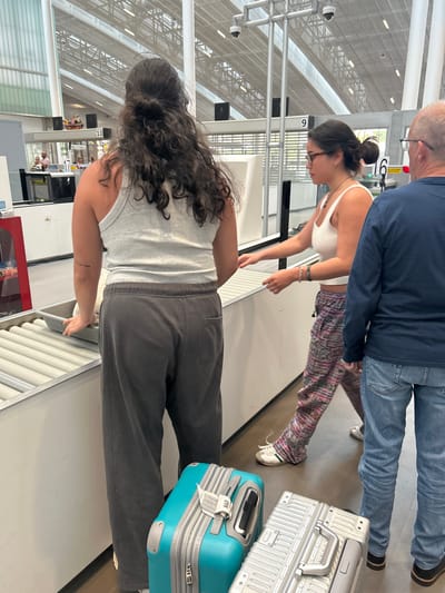 The image shows a security checkpoint at the airport in Granadilla, Spain (latitude: 28.047, longitude: -16.58). A young woman in a white tank top and pink patterned pants is placing her bag on the conveyor belt. She is talking to another woman with long dark brown hair, who is wearing a gray tank top and gray pants. She is standing next to her, looking at the conveyor belt. An older man in a gray sweater is standing behind the women, watching. The checkpoint is busy with people waiting to go through security. The environment is bright and well-lit. The floor is tiled, and there are large windows with views of the outside. There are security cameras mounted on the ceiling. There are signs indicating the numbers for each security line. There are white bins on the ground, and luggage carts next to the security lines. There are three suitcases in the foreground, one turquoise and two silver. The image suggests a normal scene of people going through airport security.